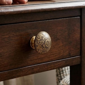 Puede incluir: Un pomo de cajón redondo de metal en tono latón con un intrincado patrón floral en relieve y detalles oscuros en bajorrelieve. El pomo está montado en el frente de un cajón de madera marrón oscuro, mostrando un acabado metálico suave.