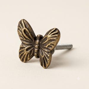 May include: A butterfly-shaped cabinet knob made of antique brass-toned metal. The surface features raised, textured wing details with dark recessed grooves. A silver-colored threaded metal bolt extends from the back of the butterfly figure.