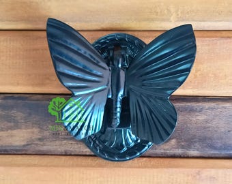 Puede incluir: Un aldaba de puerta de metal de color verde oscuro con forma de mariposa. La mariposa tiene patrones de alas detallados y está montada sobre una base circular. La aldaba está fijada a una puerta de madera.