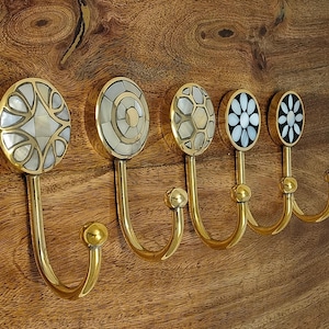 Könnte beinhalten: Fünf Wandhaken aus Messing, montiert auf einem Holzbrett. Jeder Haken hat einen runden, dekorativen Kopf, der mit weißem Perlmutt und dunklen Steinelementen in verschiedenen geometrischen, floralen und hexagonalen Mustern eingelegt ist.