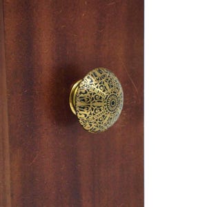 Puede incluir: Un pomo de mueble redondo con acabado en oro metálico y un intrincado patrón de filigrana negra. El pomo está montado sobre una superficie de madera de color marrón oscuro, mostrando un diseño circular detallado con líneas radiantes.