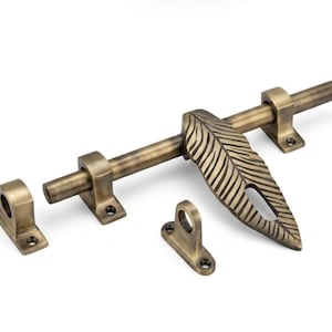 May include: A brass-toned metal door bolt featuring a sliding rod and a decorative handle shaped like a textured feather. The hardware includes two mounting brackets with screw holes and a separate strike plate, all finished in a weathered antique gold.