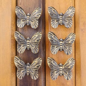 May include: A set of six butterfly-shaped knobs made of metal with a dark, antiqued bronze finish. Each knob features detailed wing patterns with raised ridges and a central body segment, arranged in two vertical columns of three on a wooden surface.