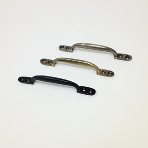 Op de afbeelding: Drie metalen ladehandgrepen gerangschikt in een diagonale rij. Elke handgreep heeft een gebogen greep en twee platte montageplaten met voorgeboorde schroefgaten. De afwerkingen omvatten matzwart, geborsteld antiek messing en een zilveren tint.