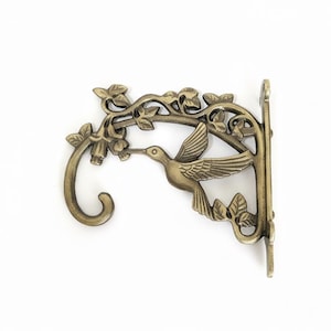 Peut inclure: Support de plante en métal couleur bronze avec un colibri. Le support décoratif représente un colibri en vol, avec des motifs floraux et de feuilles. Le crochet est incurvé et la plaque de montage comporte deux trous pour les vis. Un ajout charmant pour tout jardin.