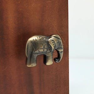 May include: A metallic elephant-shaped cabinet knob with a polished brass finish, featuring engraved details on the body and trunk. The knob is mounted on a flat, rectangular, dark brown wooden surface with a visible grain and texture.