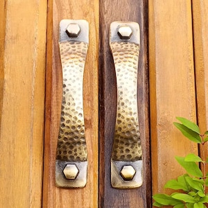 May include: Two hammered brass door pulls with a textured surface and hexagonal bolt heads. The pulls have a bronze and gold finish, and are mounted on a wooden door. The door has vertical wooden panels.