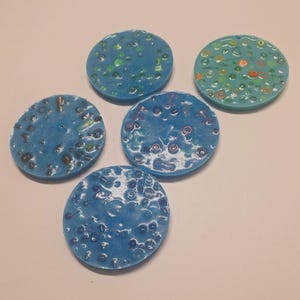 May include: Five round, blue decorative coasters with textured surfaces. Each coaster has a unique pattern of embedded colorful elements, including green, orange, and brown. The coasters are arranged on a light-colored surface.