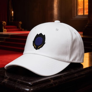 May include: White baseball cap with a dark blue and gold emblem. The cap is made of a soft material and has a curved brim. The cap is sitting on a dark surface in a room with a red carpet.