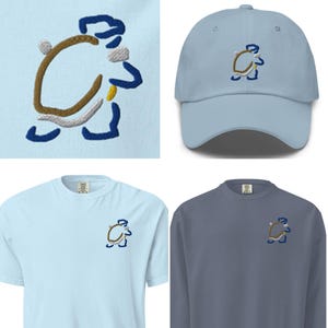May include: A light blue baseball cap, short-sleeve t-shirt, and long-sleeve sweatshirt, each featuring an embroidered cartoon design. The design is a brown, white, and blue figure. The items are displayed on a light blue background.