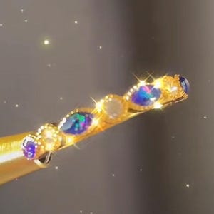 May include: A gold-toned hair accessory with a series of iridescent blue and white gemstones. The gemstones are set in a decorative pattern along the length of the accessory. The background is a soft, blurred gradient.