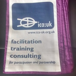 Pode incluir: Tecido roxo dobrado com um patch retangular branco com o texto "ica:uk" e as palavras "facilitation training consulting" em azul escuro e preto. O endereço do site também é visível.
