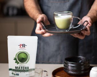 Può includere: Una busta bianca con il testo "PR- Premium Grade" e "Organic Matcha" su di essa. La busta contiene polvere di matcha verde. Una tazza di vetro di tè matcha verde è su un vassoio nero. Una ciotola di ceramica nera e un vassoio di legno con una frusta di bambù sono su una superficie bianca.