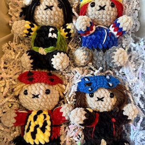 May include: Four handmade crochet plush dolls with round beige heads, black yarn hair, and embroidered facial features. Each doll wears a unique colorful outfit and headgear, including hats and masks in shades of red, blue, yellow, green, and black.