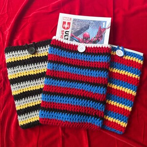 Pode incluir: Três capas de livros de crochê feitas à mão em listras pretas, amarelas, cinzas, vermelhas, azuis e brancas. Cada capa tem um fecho de botão. Uma banda desenhada com uma ilustração do Homem-Aranha é parcialmente visível.