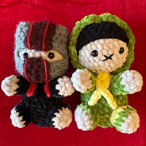 Peut inclure: Deux poupées en crochet faites à la main. La poupée de gauche est grise et noire avec des accents rouges et un visage masqué. La poupée de droite est blanche avec une tenue à capuche verte, un bandeau noir et un détail jaune en croix.