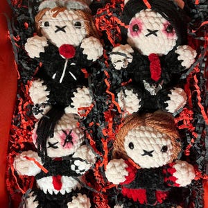 May include: Four handmade crochet dolls with black outfits, white faces, and X-shaped eyes. Each doll has unique hair and accessories, including glasses, a red lollipop, and a bat-shaped pendant. The dolls are nestled in a red box with black and red paper filler.