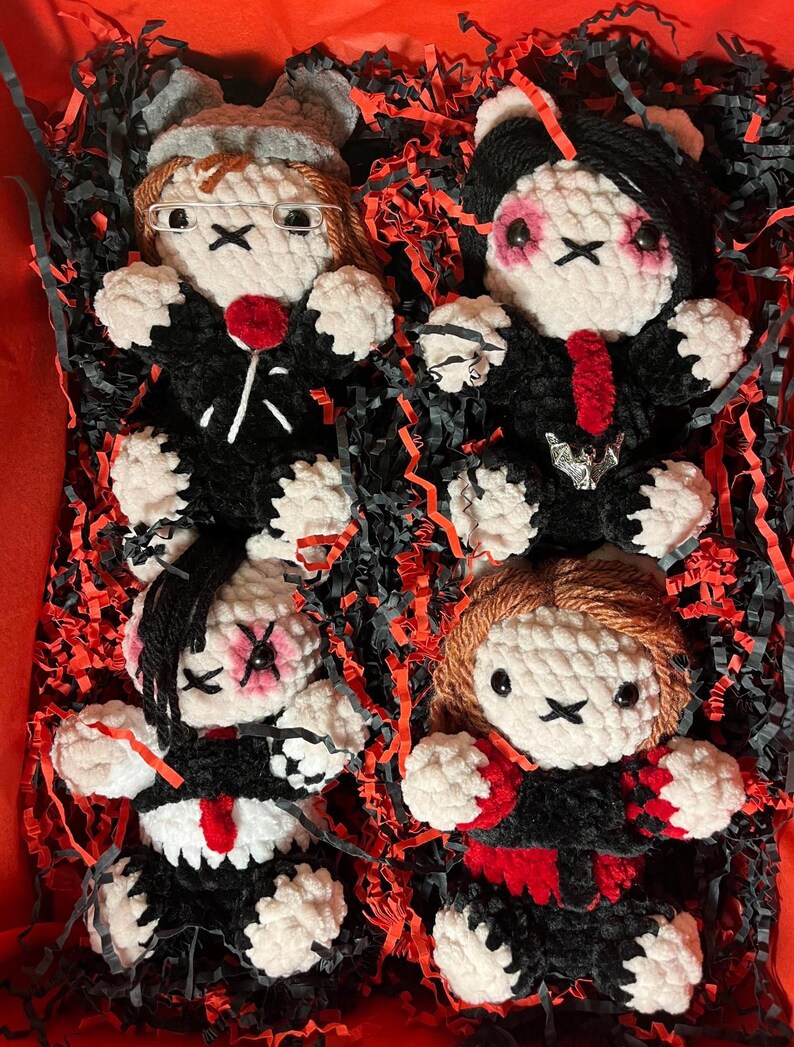 May include: Four handmade crochet dolls with black outfits, white faces, and X-shaped eyes. Each doll has unique hair and accessories, including glasses, a red lollipop, and a bat-shaped pendant. The dolls are nestled in a red box with black and red paper filler.