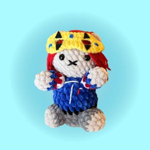 May include: A crocheted plush doll with a light gray head, red hair, and a yellow headband featuring black triangular accents. The doll wears a blue sweater with white and red sleeve details, and has gray feet. The face has black eyes and a stitched mouth.