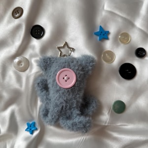 May include: A soft, grey teddy bear keyring with a pink button centre. Various buttons in shades of brown, black, and white are scattered around, along with two blue star-shaped buttons. A silver star-shaped keyring is attached.