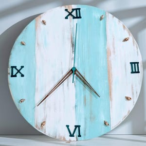 May include: A round, wooden wall clock with a distressed, painted finish. The clock face features alternating vertical stripes of white and light blue. Black Roman numerals mark the hours, and small seashells are affixed around the perimeter. The clock hands are brown.