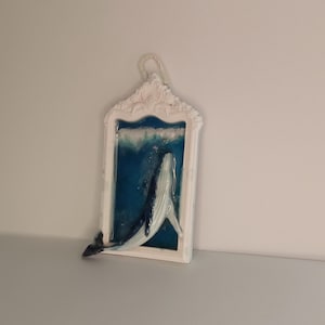 Könnte beinhalten: Weiß gerahmtes Wandbild mit einem Wal in blauem Wasser. Der Wal ist weiß und dunkelblau. Der Rahmen hat ein verziertes Design und eine Schlaufe zum Aufhängen. Wahrscheinlich aus Kunstharz.
