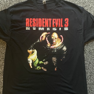 Pode incluir: Camiseta preta com o texto "RESIDENT EVIL 3 NEMESIS" em vermelho e branco. O gráfico mostra um monstro com uma arma e uma pessoa com uma camisa amarela e calças verdes. A camiseta é feita de material macio.