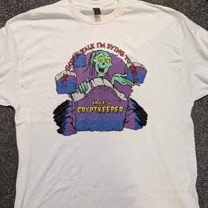 May include: White t-shirt featuring a cartoon Cryptkeeper character emerging from a tombstone. The tombstone has the text "I've got a tale I'm dying to tell" and "Tales Cryptkeeper". The Cryptkeeper is green with yellow eyes.