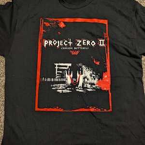 May include: Black t-shirt with a red and white graphic. The design features the text "PROJECT ZERO II" and "CRIMSON BUTTERFLY". The graphic also includes a stylized architectural scene.