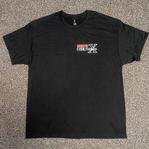 Può includere: Una t-shirt nera a maniche corte con una piccola grafica sul lato sinistro del petto. La grafica mostra il testo "RESIDENT EVIL CODE: Veronica" in bianco e rosso, accompagnato da un grande simbolo "X" bianco posizionato a destra.