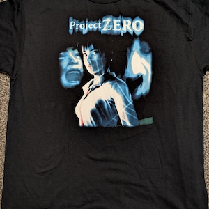 Puede incluir: Camiseta negra con un gráfico del videojuego "Project Zero". El diseño incluye un retrato de mujer con figuras espectrales azules y blancas. El texto "Project ZERO" está impreso en blanco encima del retrato.