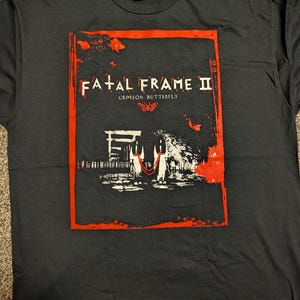 May include: Black t-shirt with a red framed graphic. The graphic features the text "FATAL FRAME II" above "CRIMSON BUTTERFLY" and a butterfly illustration. The graphic also includes a black and white image of a building.