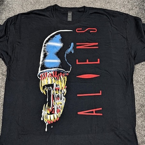Puede incluir: Camiseta negra con un gráfico de la calavera de una criatura con detalles azules y amarillos. La palabra "ALIENS" está impresa verticalmente en rojo en el lado derecho. La camiseta está hecha de un material suave.