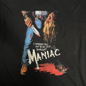 Peut inclure: T-shirt noir avec un graphisme du film d'horreur "Maniac". Le motif représente une figure tenant un couteau ensanglanté et une tête coupée, avec les mots "I warned you not to go out tonight" et "Maniac" en blanc.