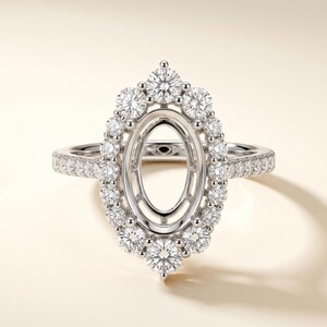 May include: A silver-toned ring featuring an empty oval-shaped center setting surrounded by a halo of small, round, clear stones. The band is also adorned with a row of small, round, clear stones along the sides, creating a sparkling effect.