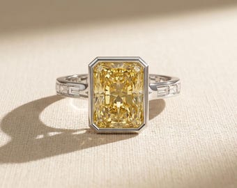 May include: A ring featuring a large rectangular radiant-cut yellow center stone set in a silver-toned metal bezel. The band is accented with small rectangular baguette-cut clear stones embedded along the sides of the metal shank.