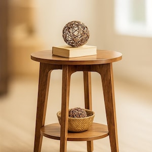 May include: A round, wooden side table with a lower shelf and three legs. A woven sphere sits atop a book on the table's surface. A woven basket on the lower shelf holds another sphere. The table is made of light brown wood.