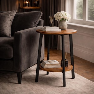 May include: A round, two-tiered side table with a dark wood top and shelf, supported by black metal legs. The table holds books, a vase of white flowers, and a black diffuser. The table is next to a gray sofa.