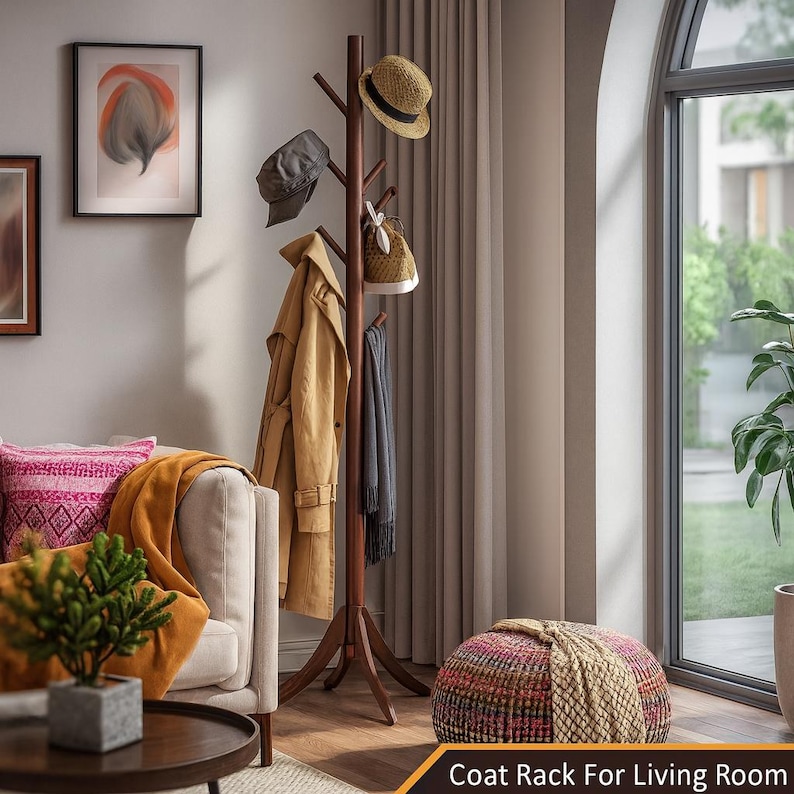 May include: A dark brown wooden coat rack with multiple hooks, holding a tan trench coat, a gray scarf, and various hats and a bag. The rack is in a living room setting with a window and a couch.