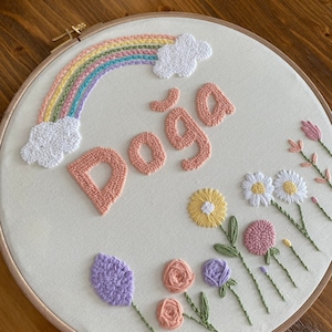 Può includere: Un cerchio da ricamo bianco con un arcobaleno, nuvole, il nome "Doga" in rosa e vari fiori ricamati in rosa, viola, giallo e verde.