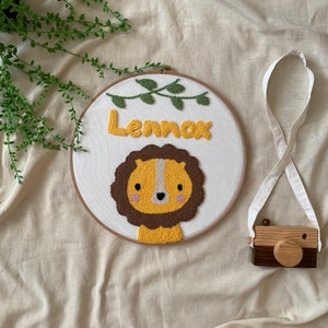 Può includere: Un cerchio da ricamo bianco con un viso di leone marrone e il nome "Lennox" ricamato con filo giallo. Una piccola macchina fotografica in legno con un cinturino marrone si trova nell'angolo in basso a destra.