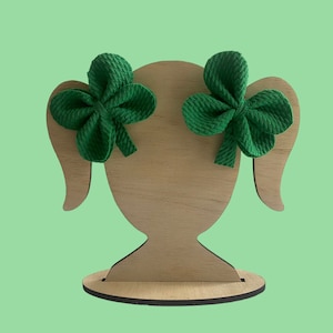 May include: Two green fabric clover-shaped hair bows displayed on a wooden head-shaped stand. The bows are a vibrant green colour and have a textured appearance. The stand is made of light-coloured wood and has a simple, stylized design.