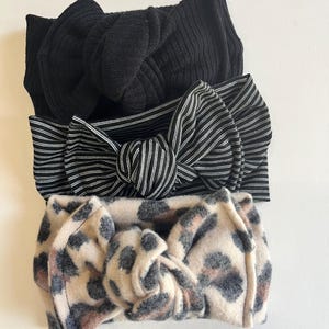 May include: Three knotted headbands stacked vertically. The top headband is solid black with a ribbed texture. The middle headband features a black and white pinstripe pattern. The bottom headband has a tan and black leopard print design.