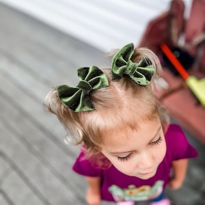 K&ouml;nnte beinhalten: Ein junges M&auml;dchen mit blonden Haaren, das ein rosa Shirt und zwei gr&uuml;ne Samt-Haarb&auml;nder tr&auml;gt.