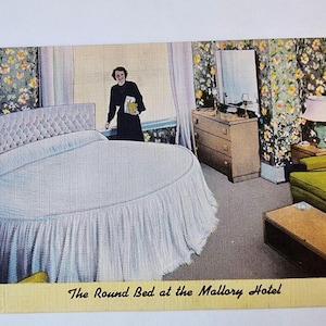 May include: Vintage postcard featuring a round bed in a hotel room. The bed has a light blue bedspread and a ruffled skirt. The room includes a dresser, sofa, and floral wallpaper. Text reads "The Round Bed at the Mallory Hotel."