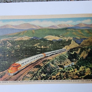 May include: Vintage postcard featuring a Santa Fe Streamliner train descending Cajon Pass on the Southern California side of the Coast Range. The train is orange and white, set against a backdrop of green hills and mountains.
