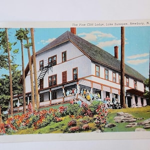 May include: Vintage postcard showing The Pine Cliff Lodge in Newbury, N.H. The white building has a dark roof, a porch, and a fire escape. Colourful flowers and trees surround the lodge, with a group of people on the porch. The text reads "The Pine Cliff Lodge, Lake Sunapee, Newbury, N.H."
