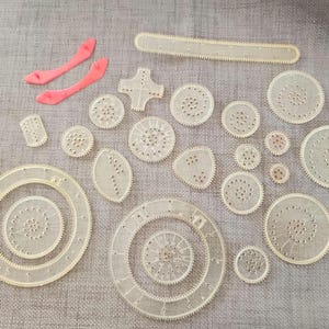 May include: A selection of clear plastic Spirograph gears and shapes, including circles, ovals, and a straight edge. Two bright pink curved pieces are also present. The objects are arranged on a grey fabric surface.