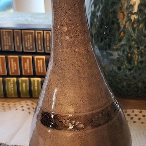 May include: A handcrafted, speckled brown ceramic vase with a flared top and a bulbous base. The vase features a dark brown band with a floral design near the base. The vase is a decorative item.