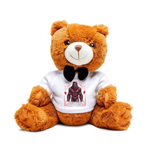 Puede incluir: Un oso de peluche marrón que lleva una camisa blanca con una pajarita negra. La camisa tiene un gráfico de un sasquatch marrón con el texto "Candy Thief" y "Beware Sweet Tooth"